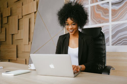 Mujer En Chaqueta Negra Sentada Junto A La Mesa Mientras Usa Macbook