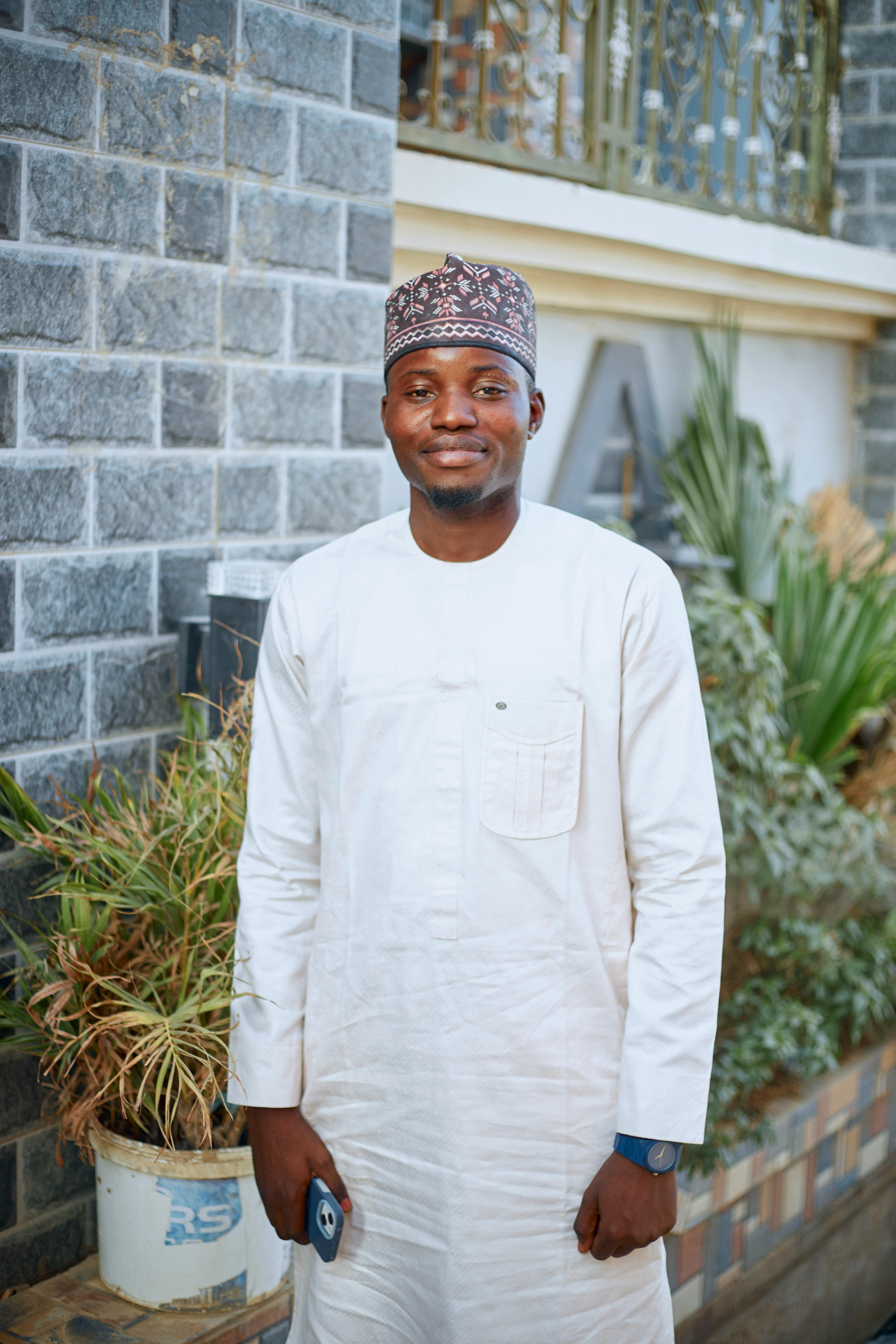Portrait of a smiling man wearing traditional attire outdoors, standing in front of a brick wall.