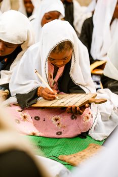Children in traditional attire writing on wooden boards during a cultural education session.