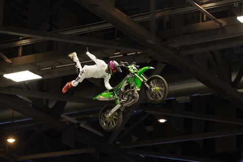 Free Thrilling indoor motocross stunt with rider mid-air above a green bike inside an arena. Stock Photo