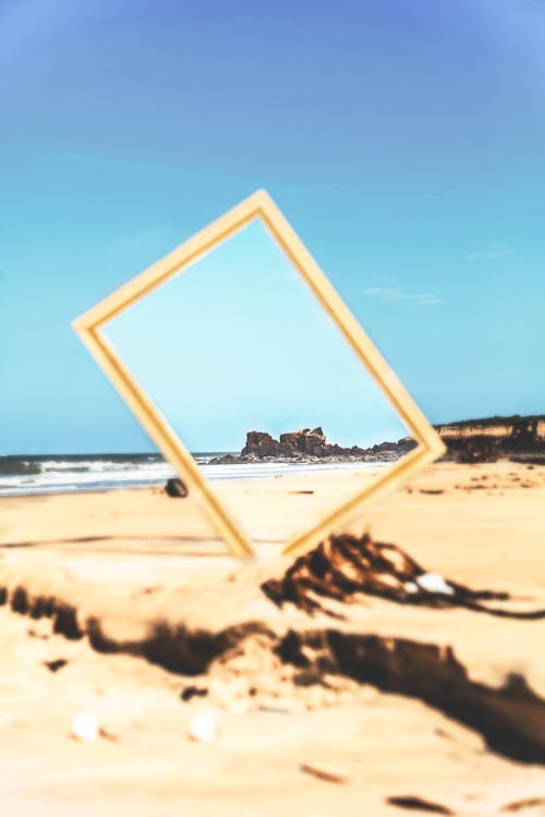 Mirror in wooden frame placed in sand on seaside on background of blue sky and rocks