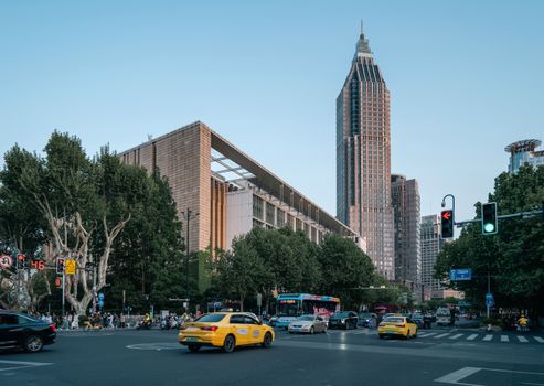 A bustling street scene near Nanjing Library with taxis and a towering skyscraper under a clear sky.