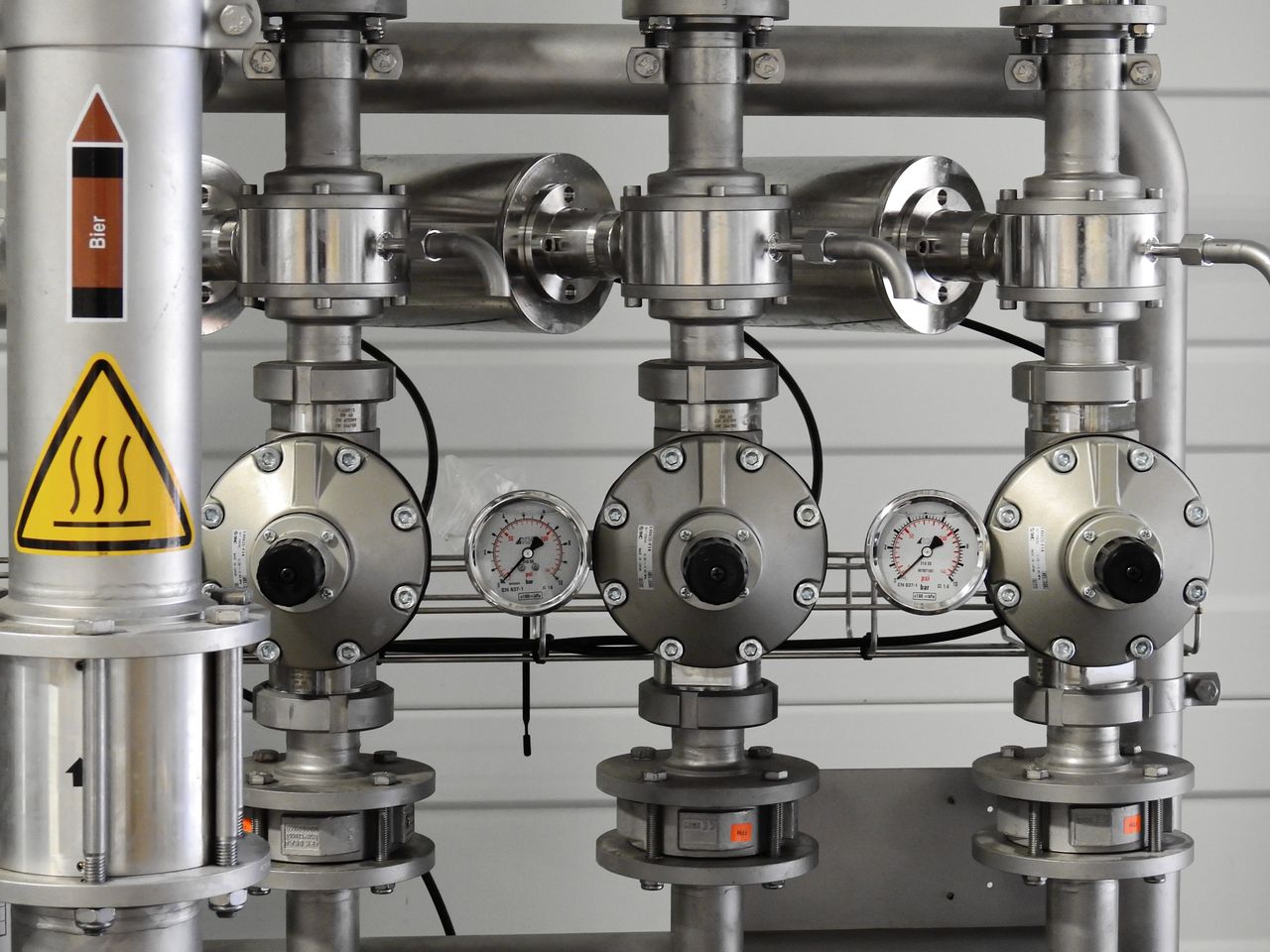 Stainless steel pipe manifold fitted with pressure gauges and isolation valves against a plant wall.