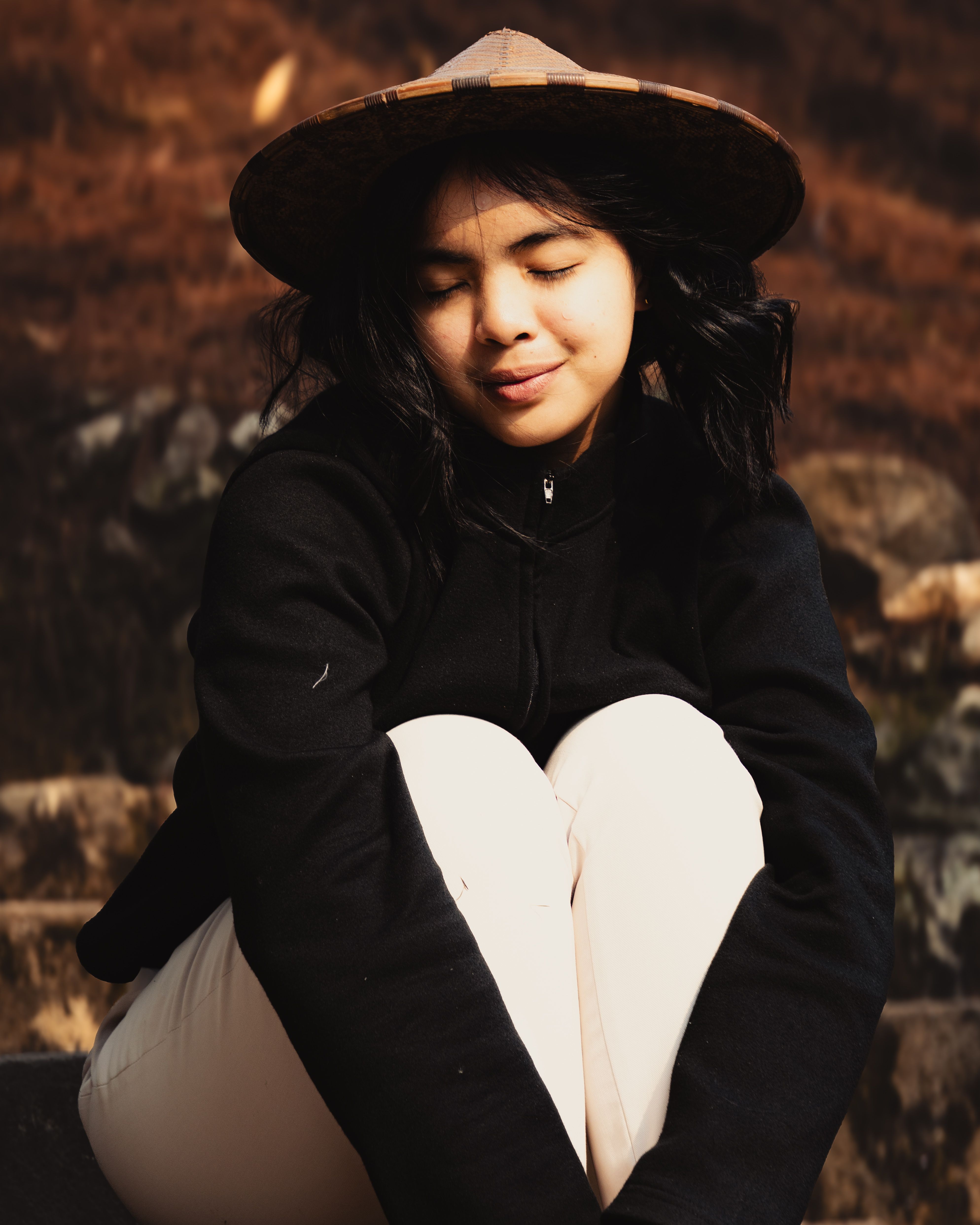 Peaceful portrait of young woman with a hat sitting outdoors, eyes closed.
