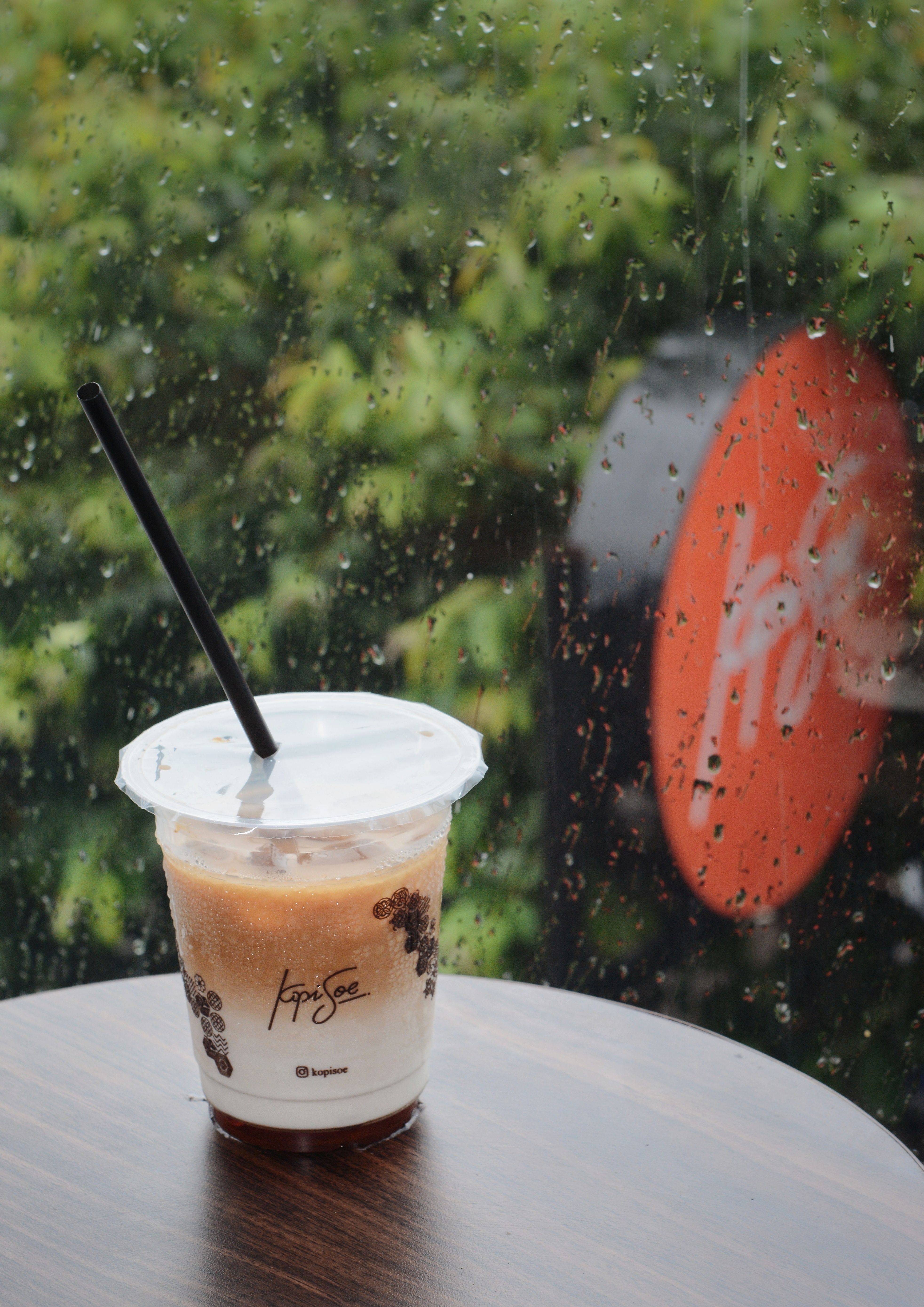 Iced coffee on table, raindrops on window at a Bandung cafe. Captures cozy and rainy ambiance.