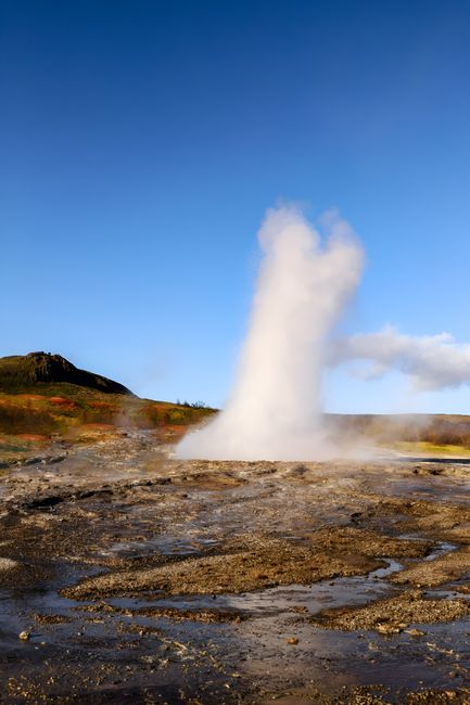 A dramatic geyser erupts on a remote island under a clear blue sky.