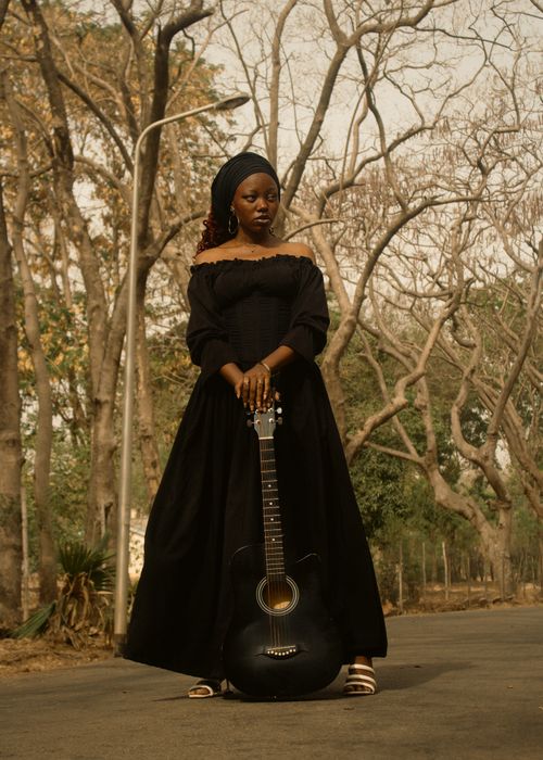 Free A woman in a black dress stands with a guitar on a tree-lined road, exuding grace and creativity. Stock Photo