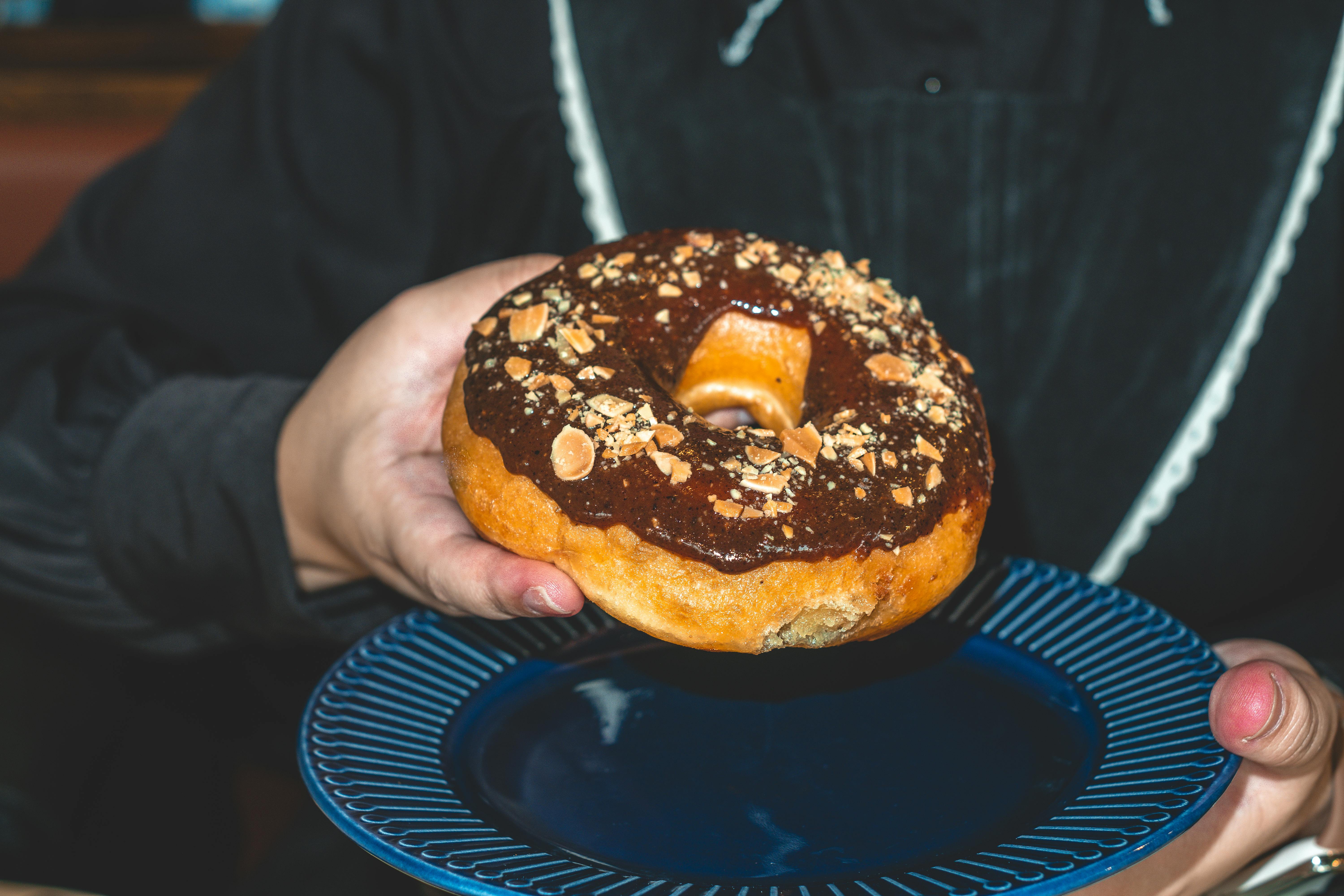Close-up of a hand holding a chocolate donut topped with nuts on a blue plate.