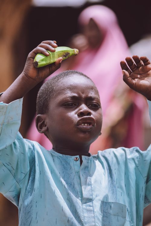 Free A young boy energetically holding a fruit with a blurred figure in the background, creating an engaging outdoor scene. Stock Photo
