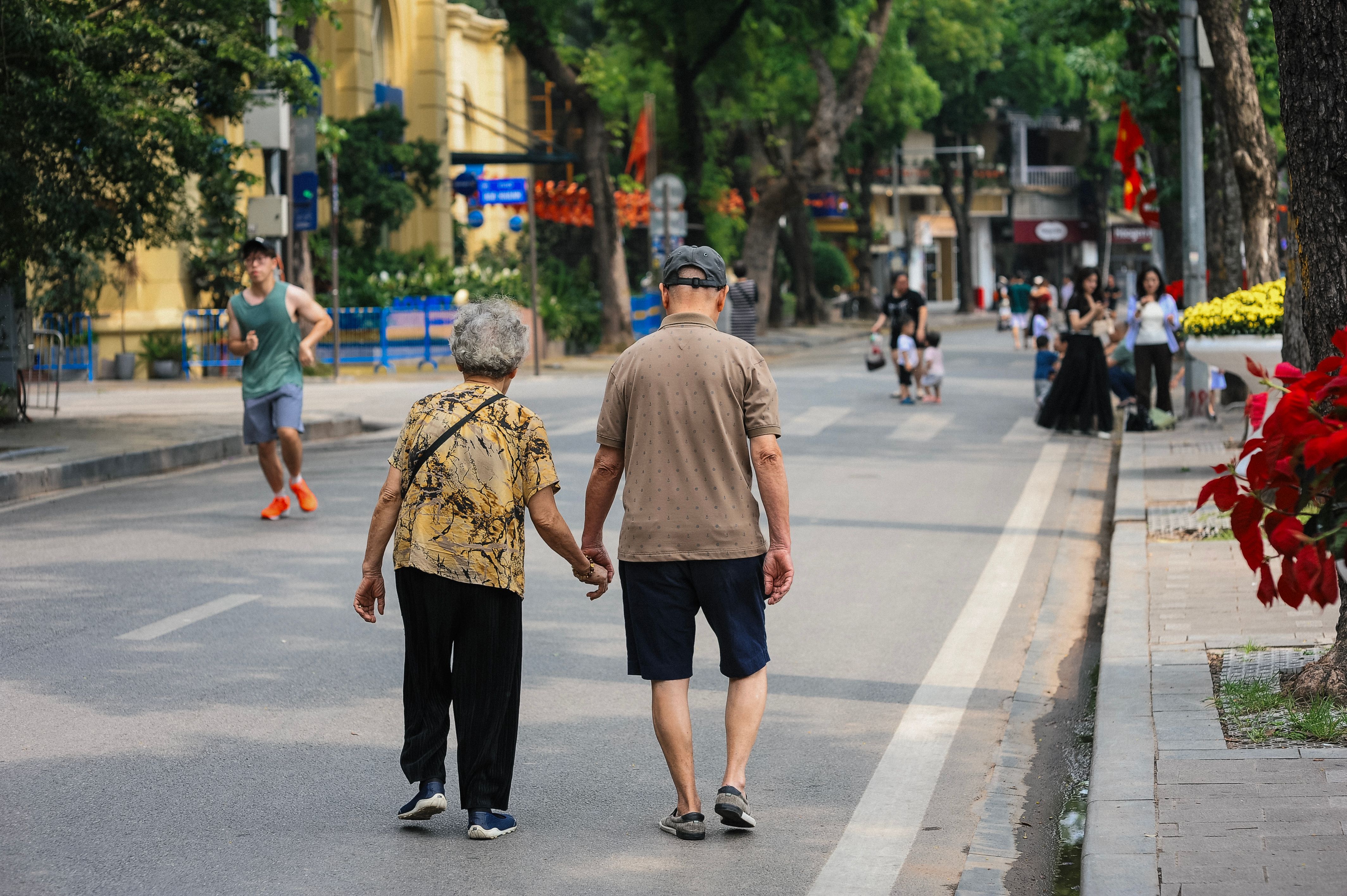 A senior couple walking hand in hand along a vibrant city street filled with people and greenery.