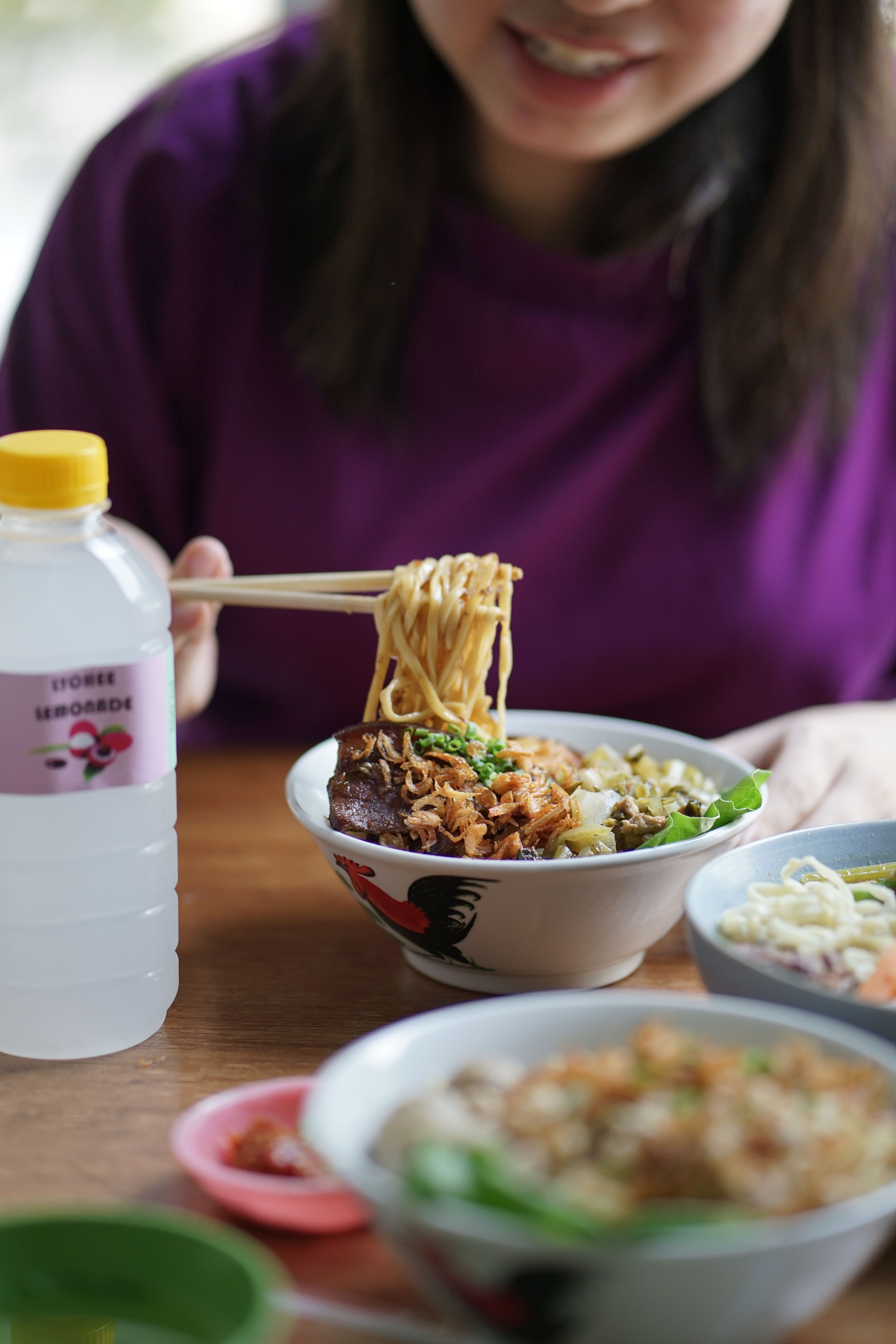 A woman in a purple shirt savoring a bowl of Asian noodles with chopsticks. Bottled drink on table.