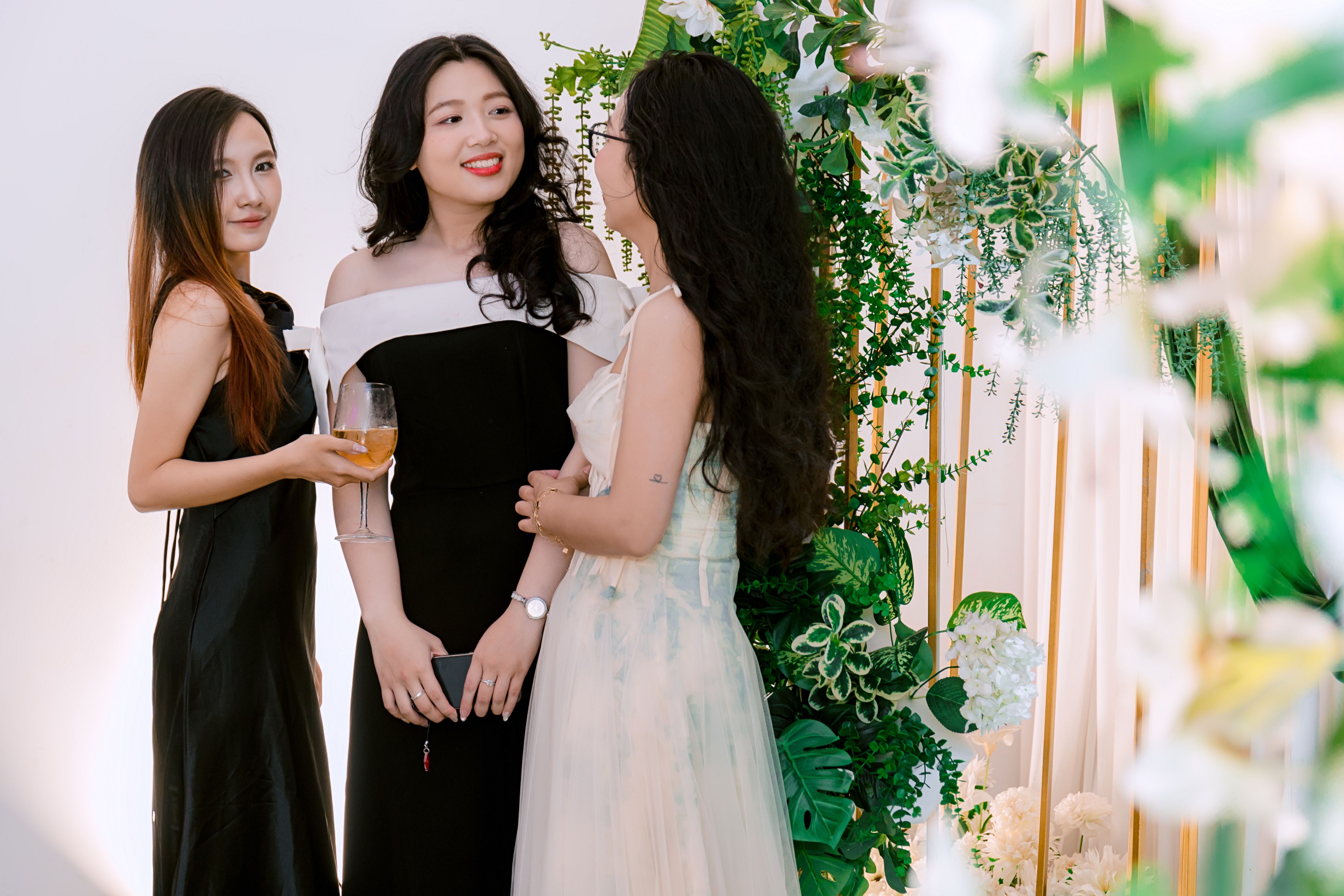 Três mulheres elegante conversando e sorrindo em um evento social decorado com plantas verdes e flores brancas.