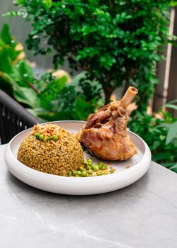 Grilled lamb shank served with freekeh and peas on a white plate outdoors in Dubai.