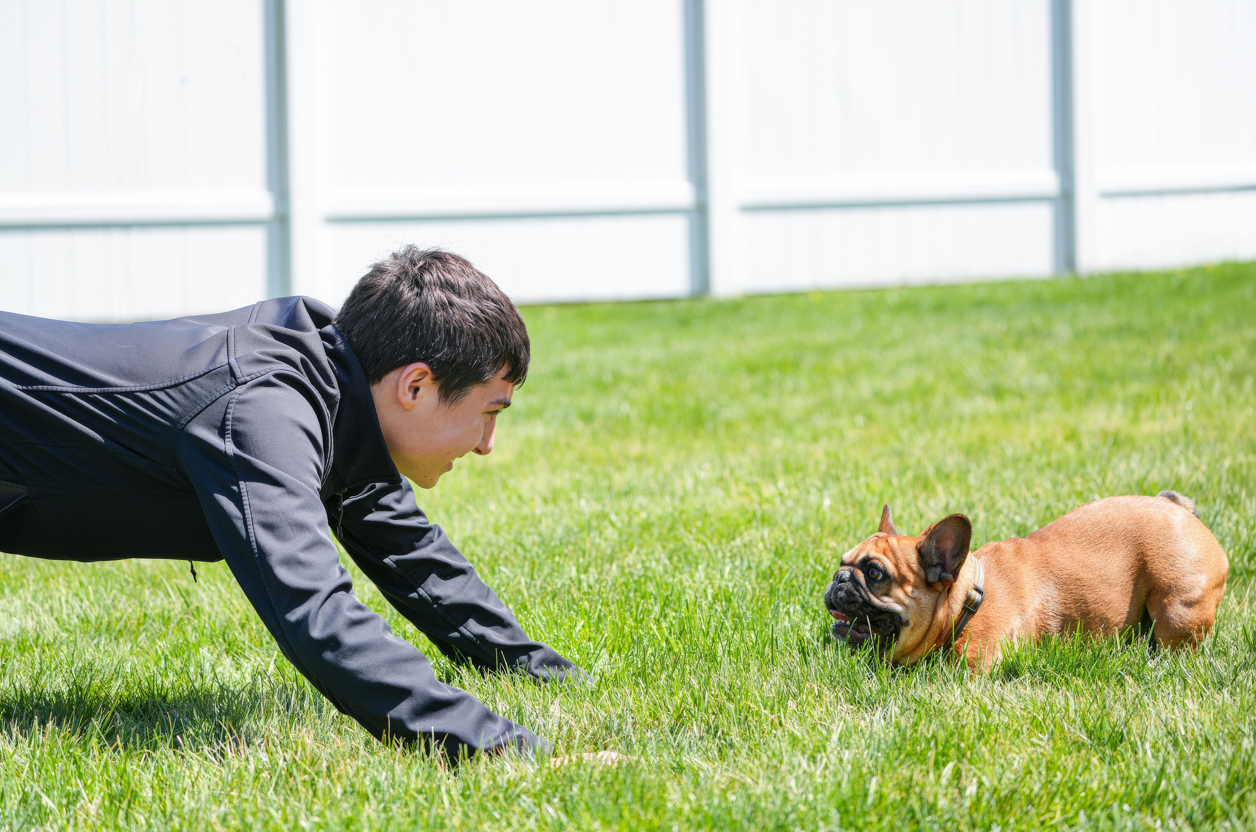 Teen engaging in play with a French Bulldog on a bright, sunny lawn outdoors.