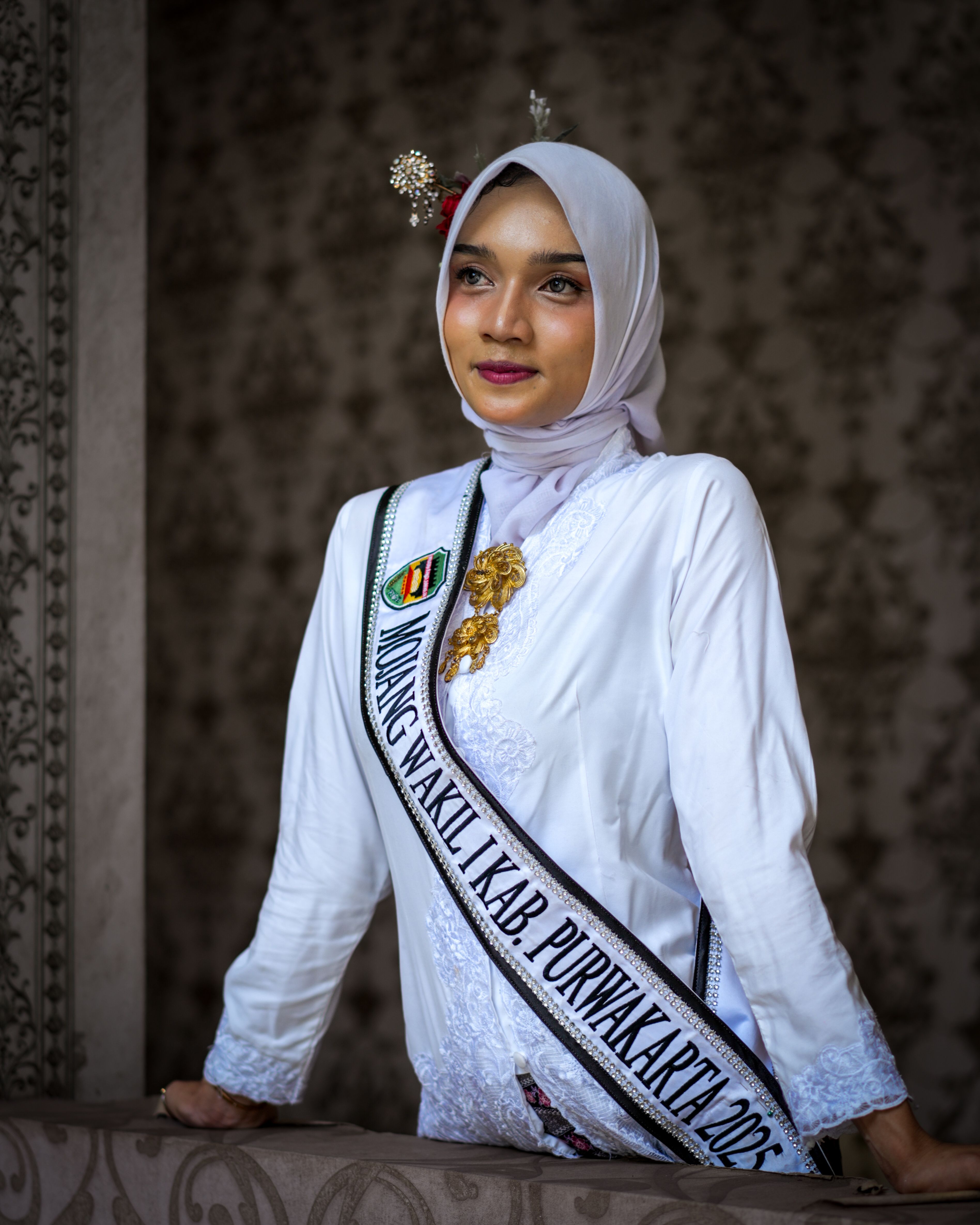 Young woman in traditional attire and sash standing indoors, looking poised and elegant.