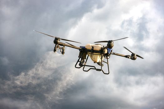 A drone dispersing mist in a cloudy sky, showcasing agricultural technology innovation.