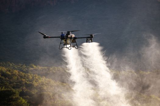 A drone disperses pesticides over a lush green farmland with mountains in the background, captured at dawn.