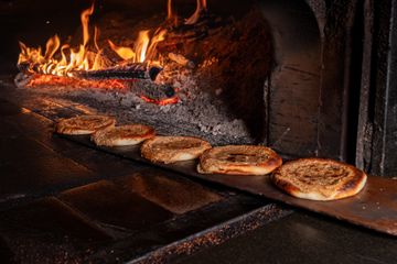 Round flatbreads lined up on a long peel beside a blazing wood-fired oven