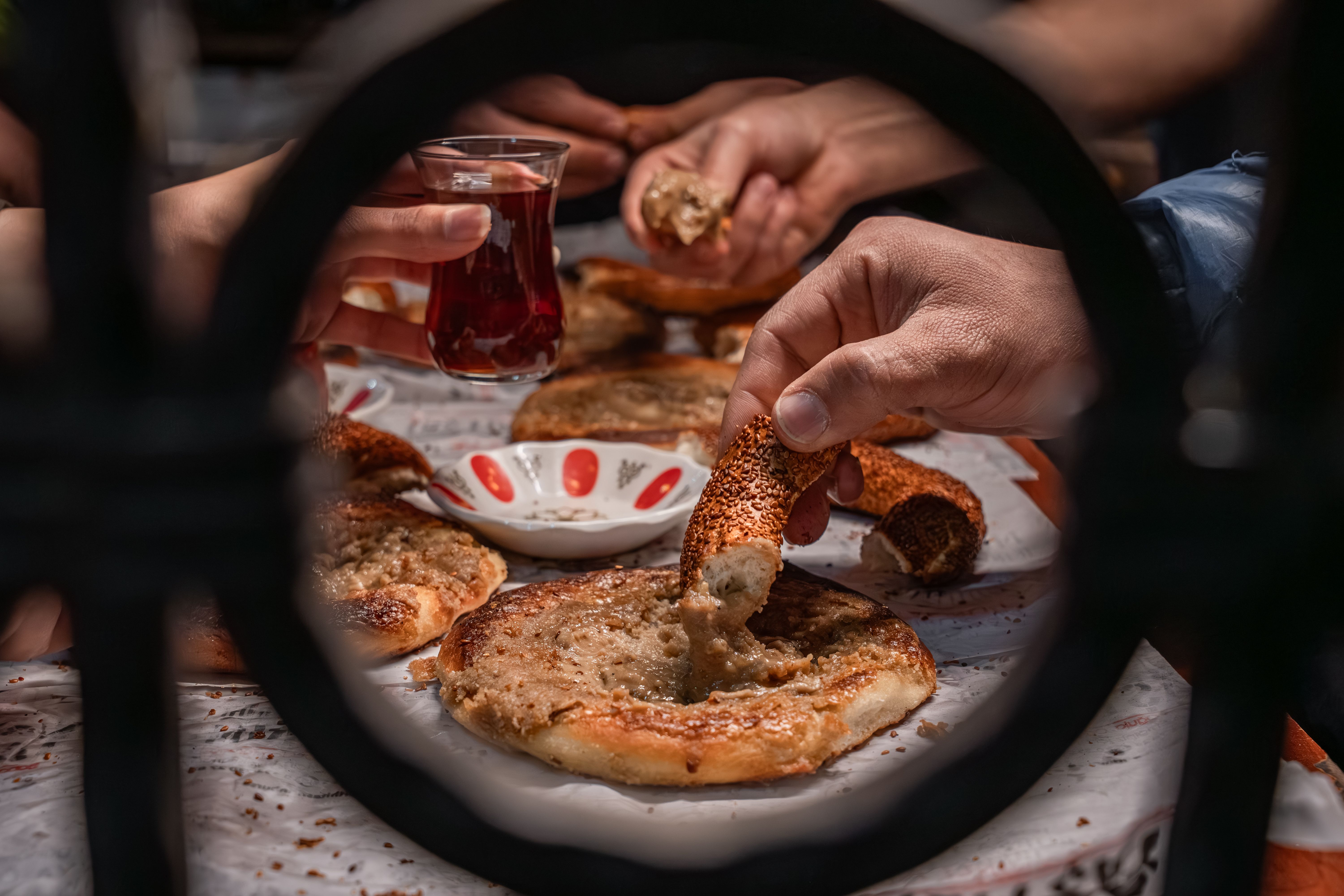 Hands sharing Turkish simit with tea during a communal breakfast, viewed through a decorative gate.