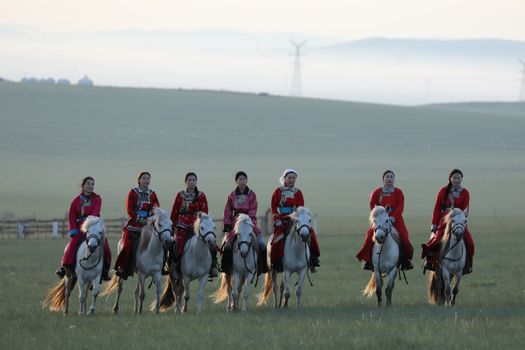 A group of Mongolian women in traditional costumes riding white horses on a grassy plain at dawn.