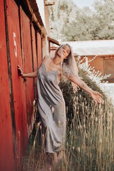 A young woman with long hair in a slip dress poses by a rustic red door surrounded by tall grass in a summer setting.