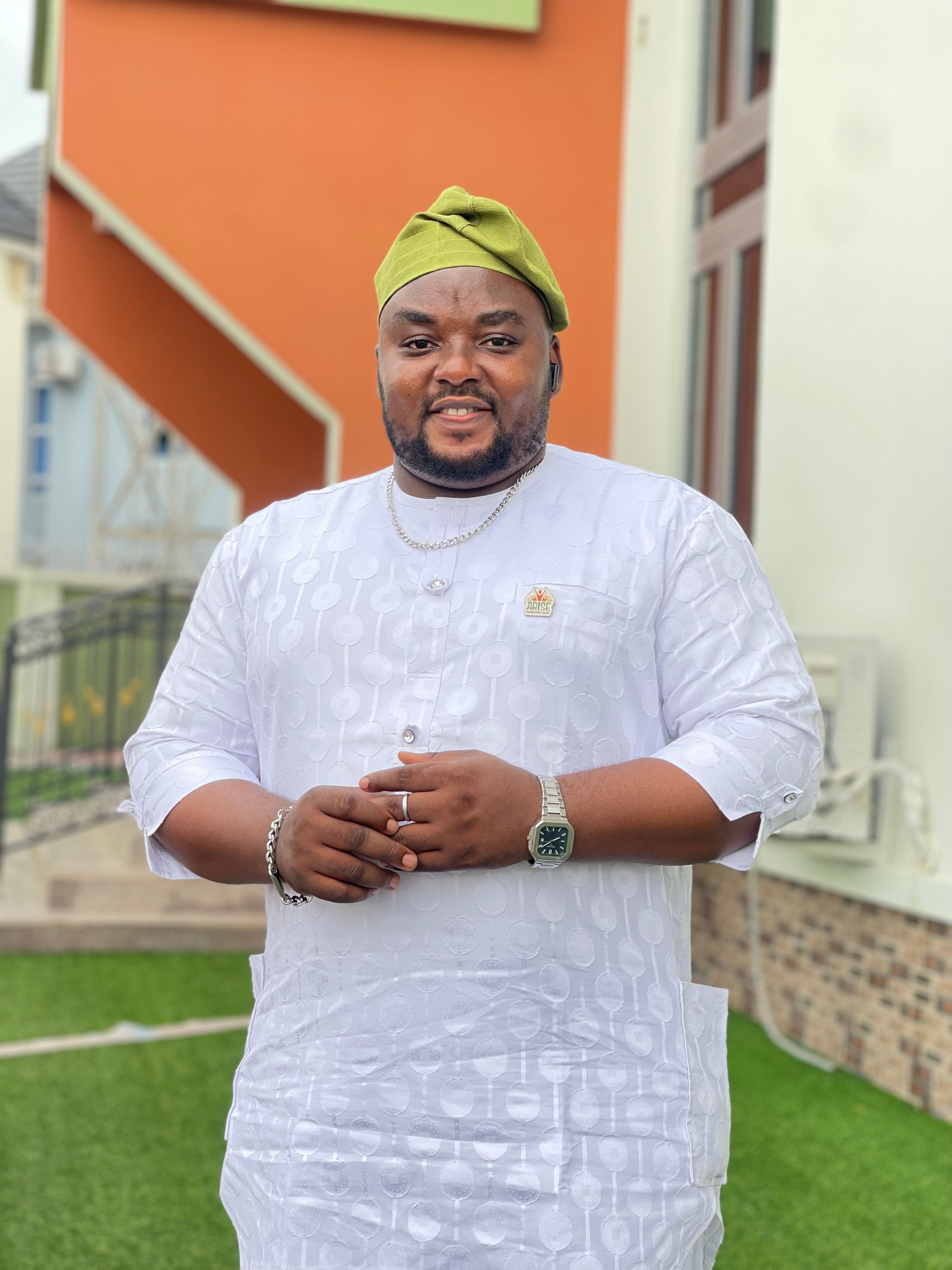 Smiling man in traditional Nigerian attire and cap, posing outdoors.