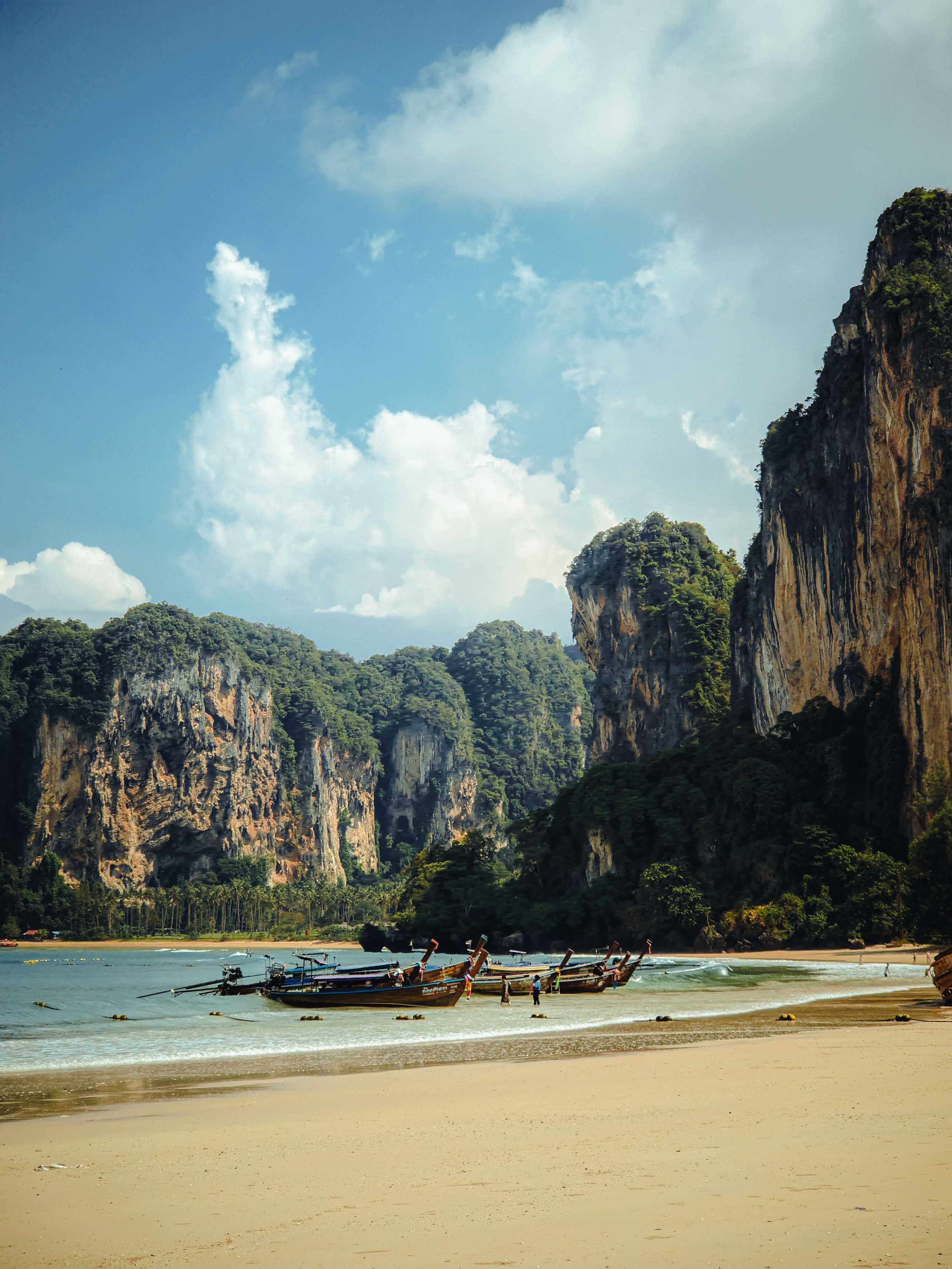 Serene beach with towering cliffs in Krabi, Thailand, showcasing traditional longtail boats under a clear sky.