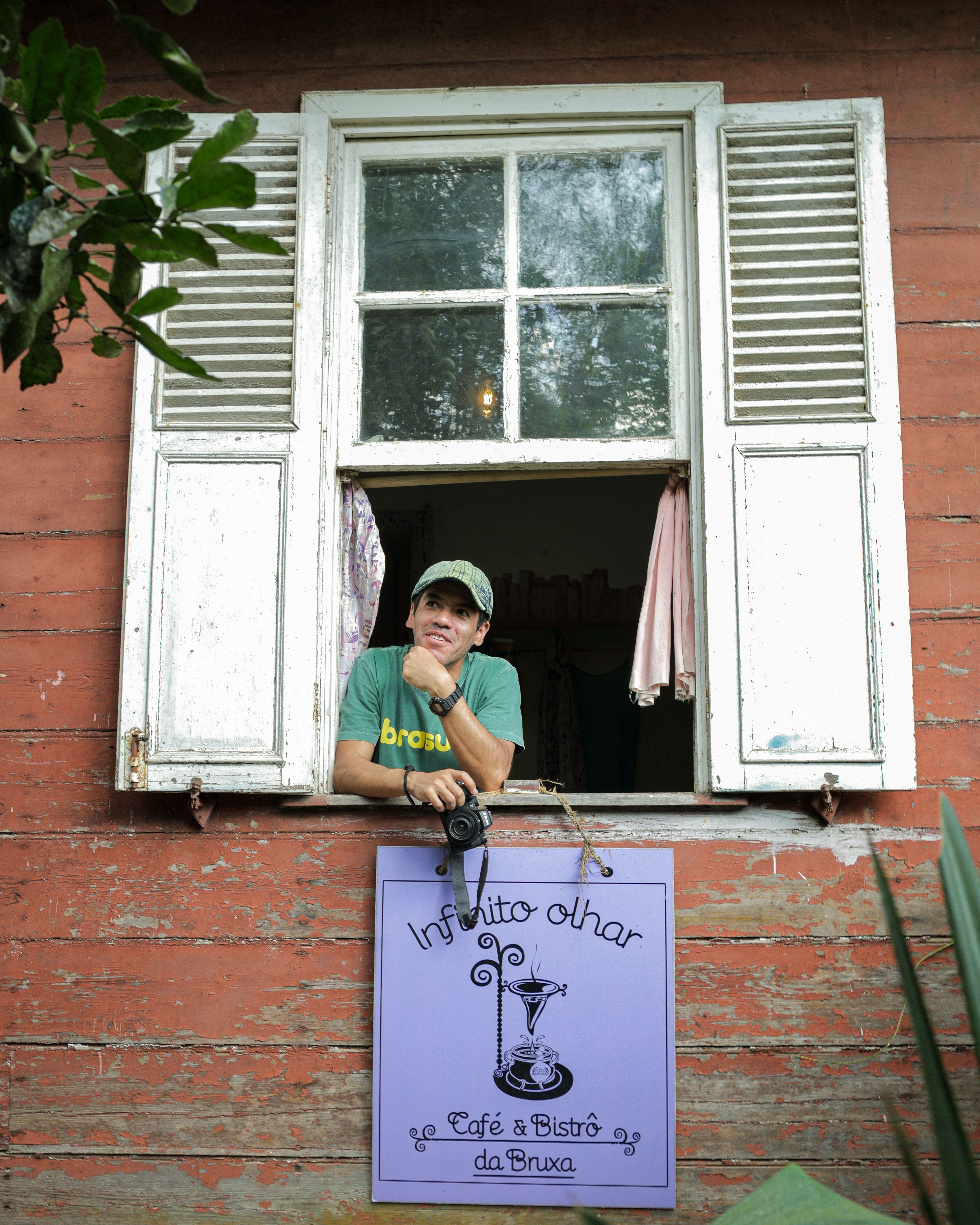 Individual at an open window with a camera, leaning on a café sign in a rustic setting.