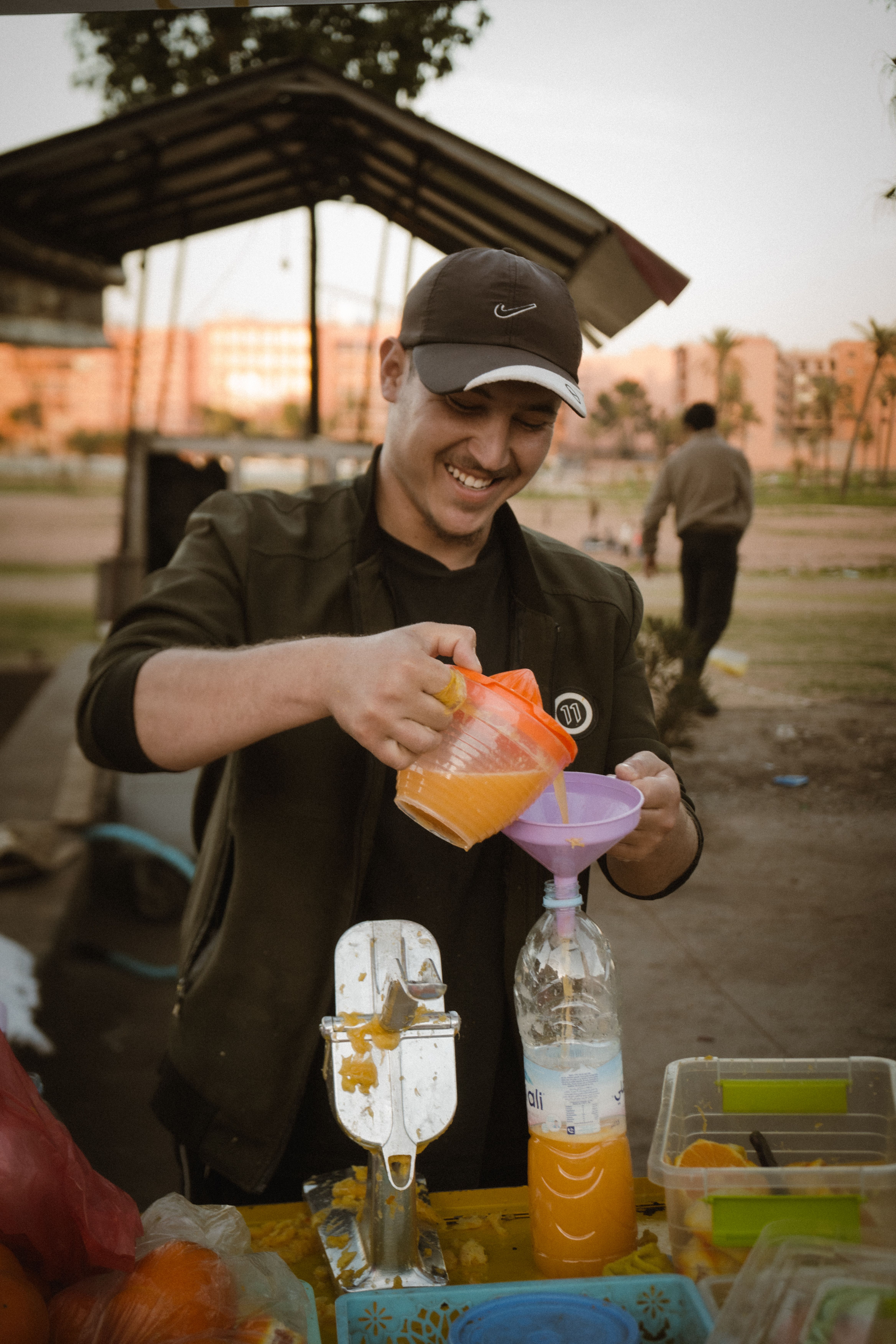 A smiling street vendor pouring fresh orange juice into a bottle in an outdoor setting.