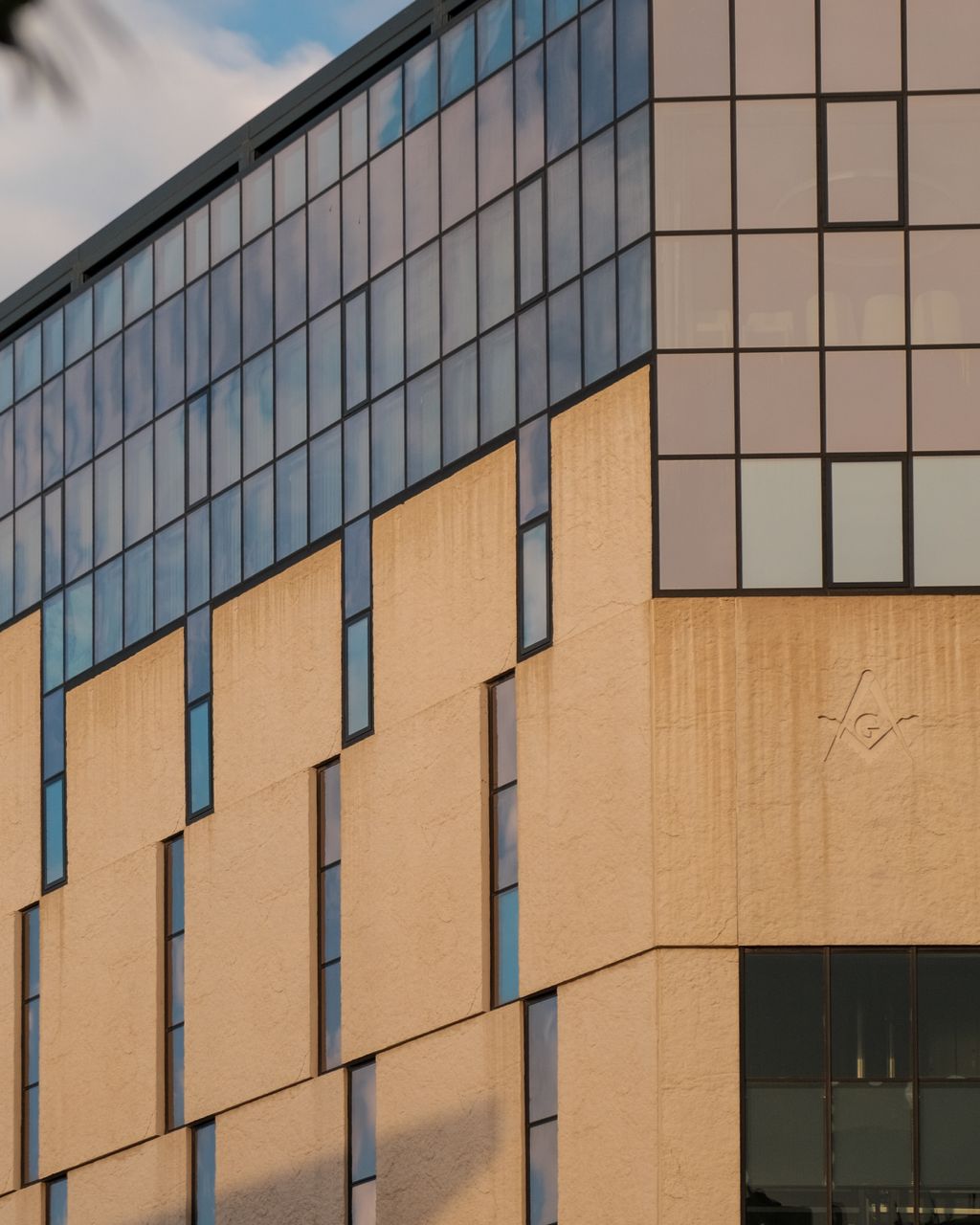 The corner of a modern commercial building at golden hour, with a pale stone lower facade of narrow vertical windows and a glazed upper storey reflecting the sky.
