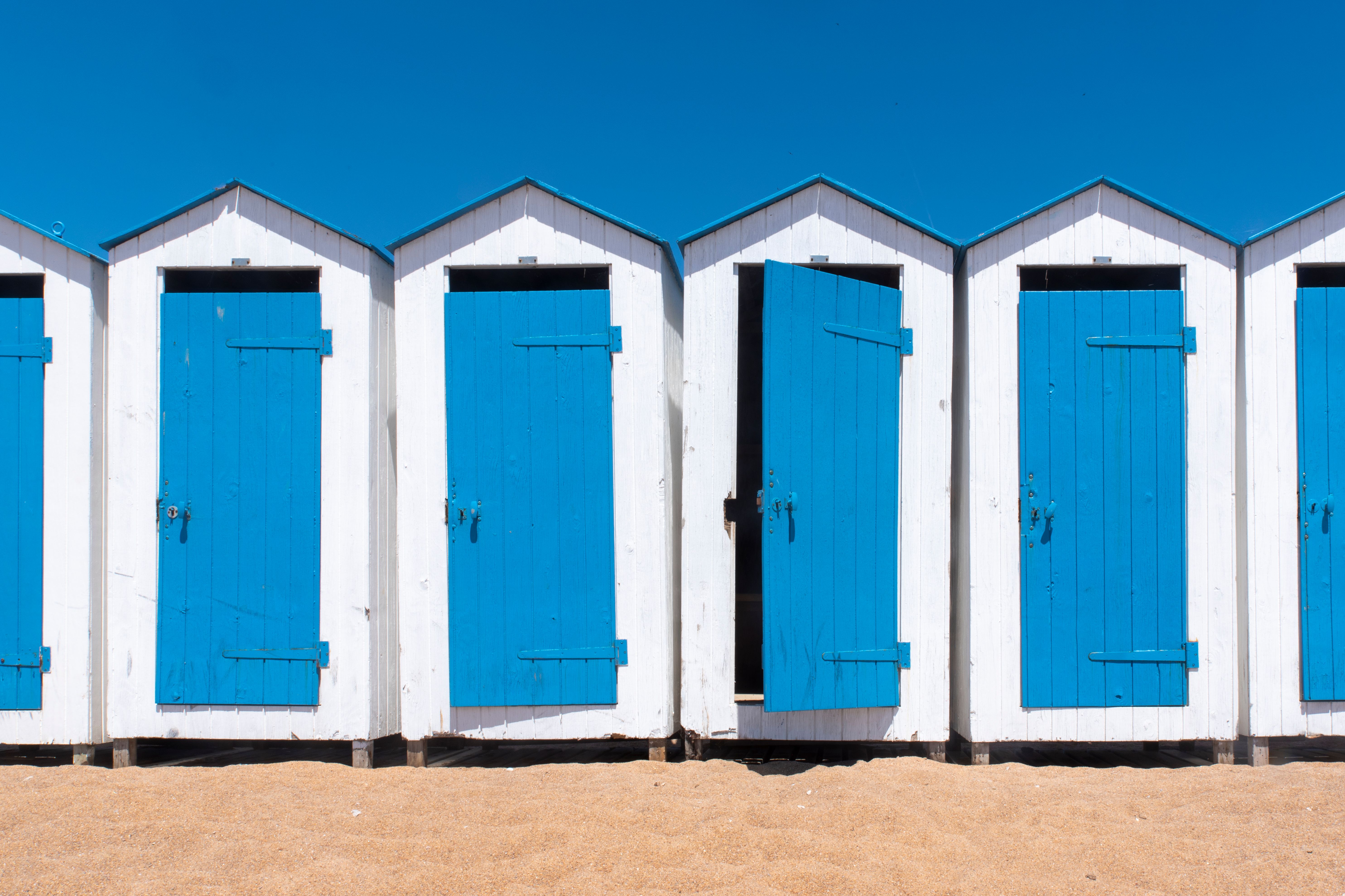 White beach cabins with blue doors on a sandy coast under a clear blue sky, conveying summer relaxation.