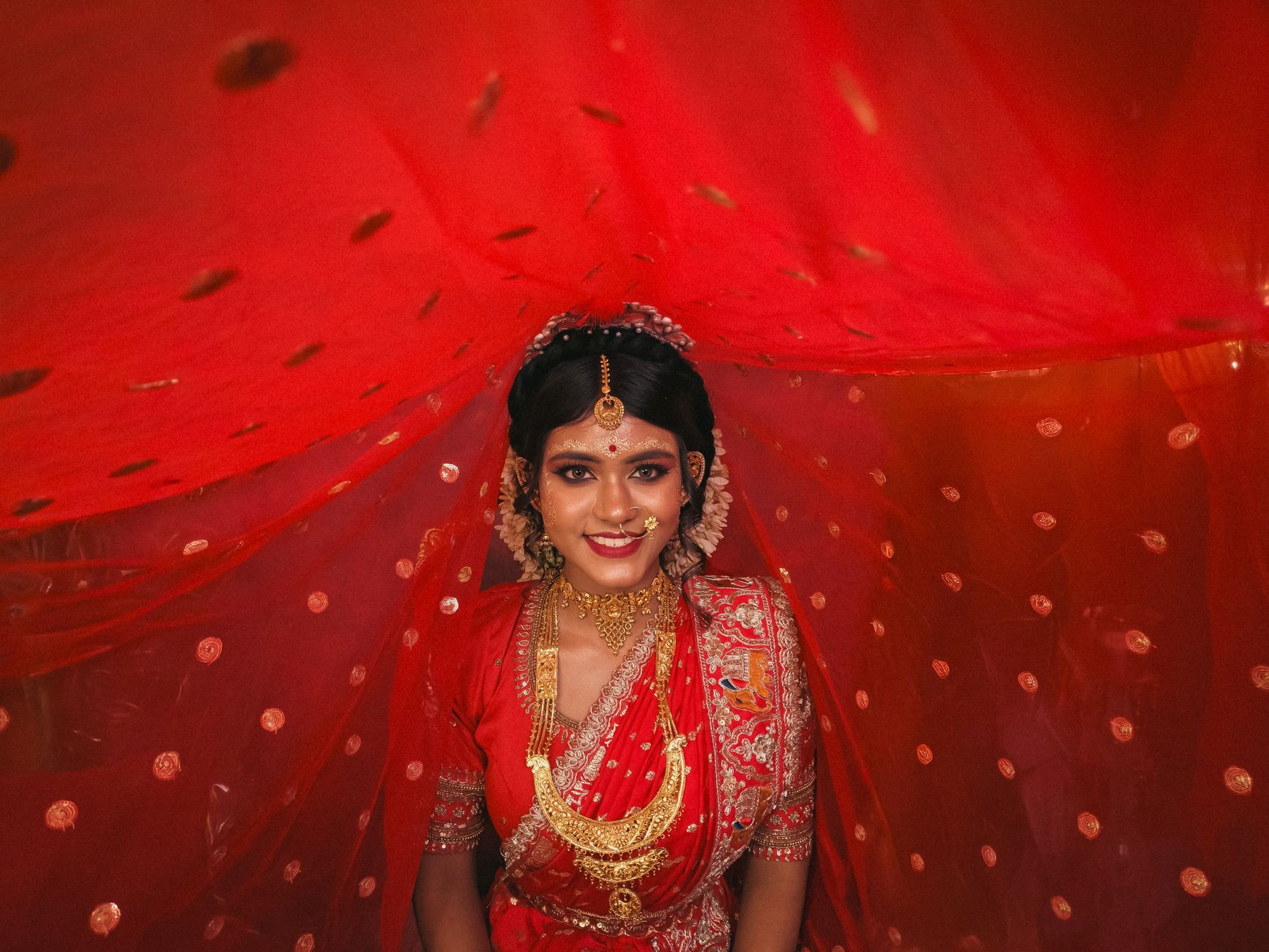 Beautiful Indian bride in red sari and gold jewelry, smiling under a red veil in Kolkata.