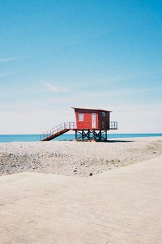A solitary red beach hut stands on a rocky shoreline under a clear blue sky, evoking a sense of isolation.