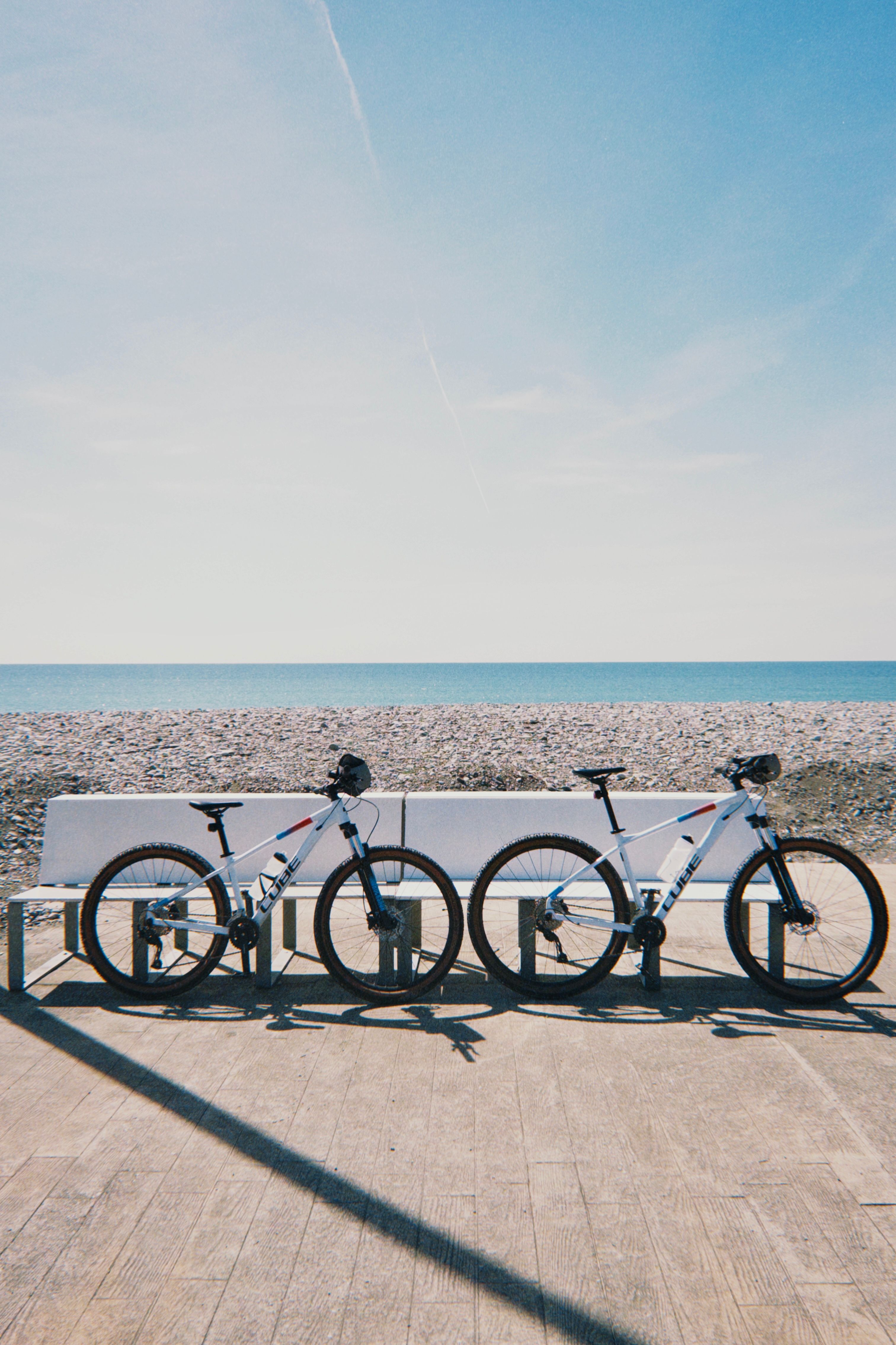 Two bicycles rest by a beach with a vast, tranquil ocean under a clear sky, capturing a serene coastal scene.