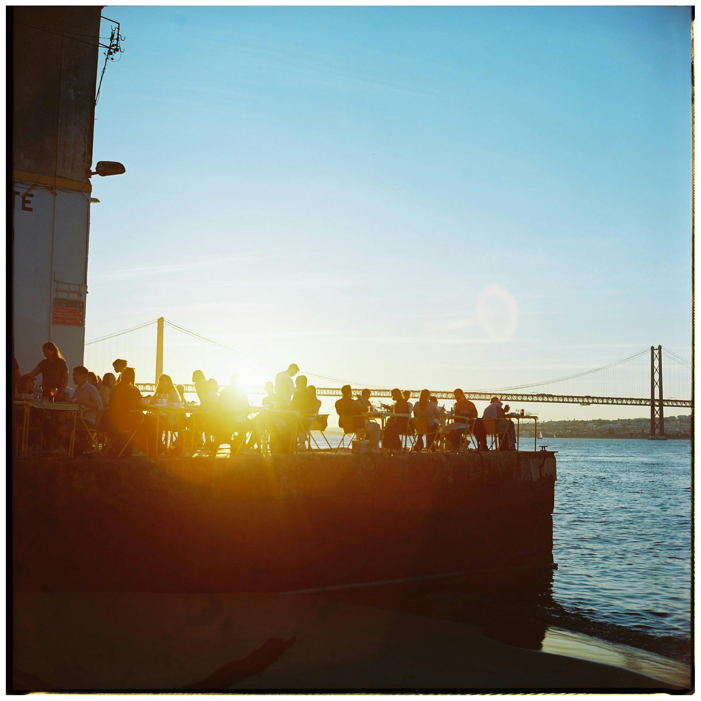 People enjoying a sunset near Lisbon's iconic 25 de Abril Bridge, capturing the vibrant city life.