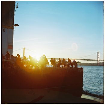 People enjoying a sunset near Lisbon's iconic 25 de Abril Bridge, capturing the vibrant city life.