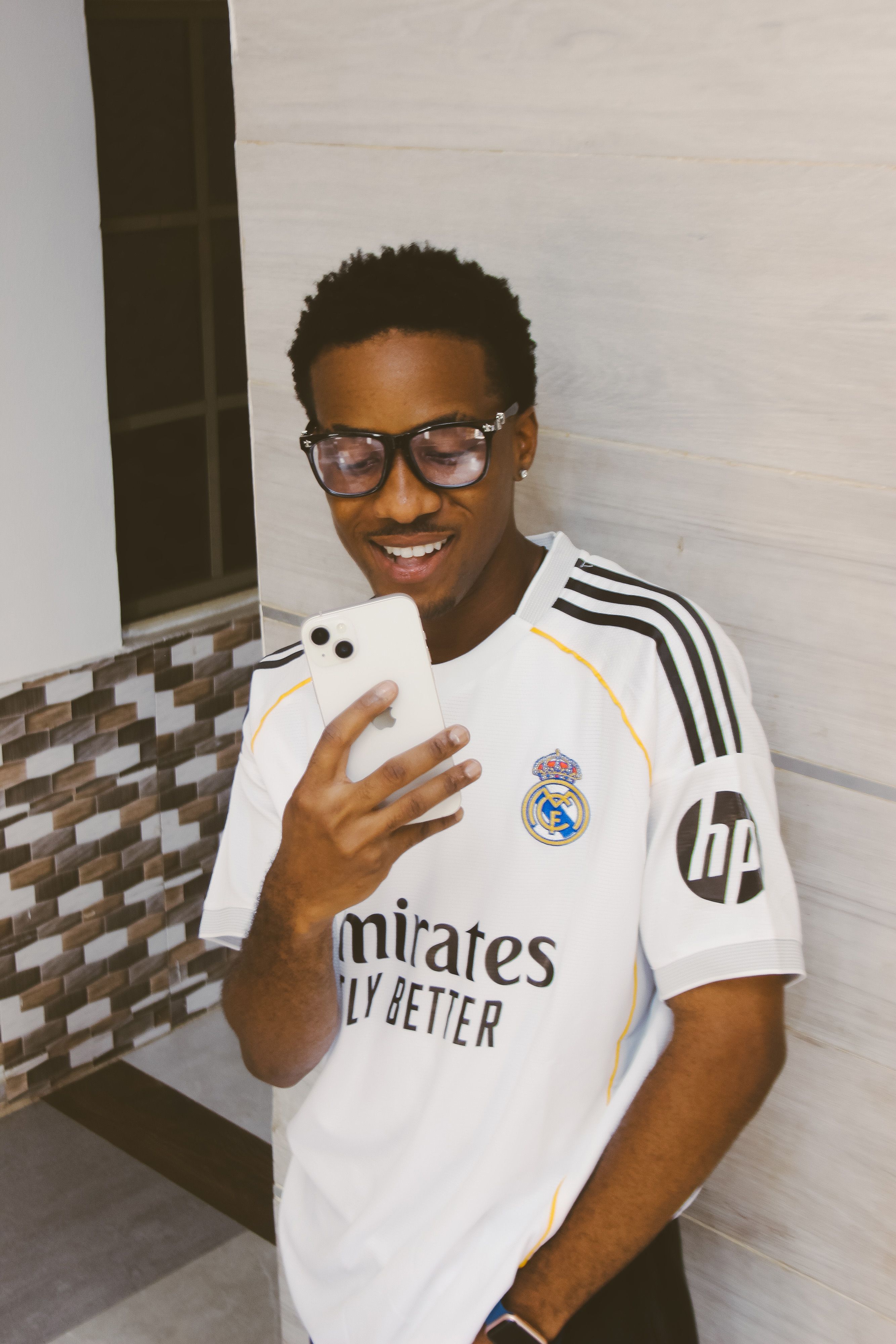 A young man in glasses smiles while using his smartphone indoors.