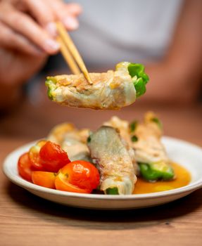 Close-up of stuffed pepper wrap held by chopsticks, showcasing Asian cuisine with cherry tomatoes.