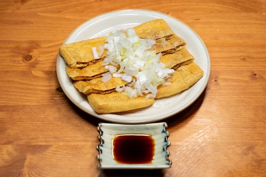 Crispy fried tofu topped with chopped onions, served with soy sauce on a wooden table.