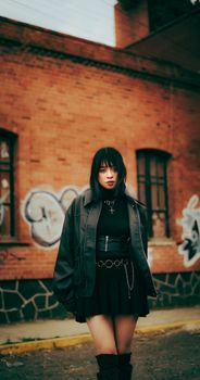 Young woman in black outfit stands confidently against graffiti brick wall. Edgy and modern vibe.