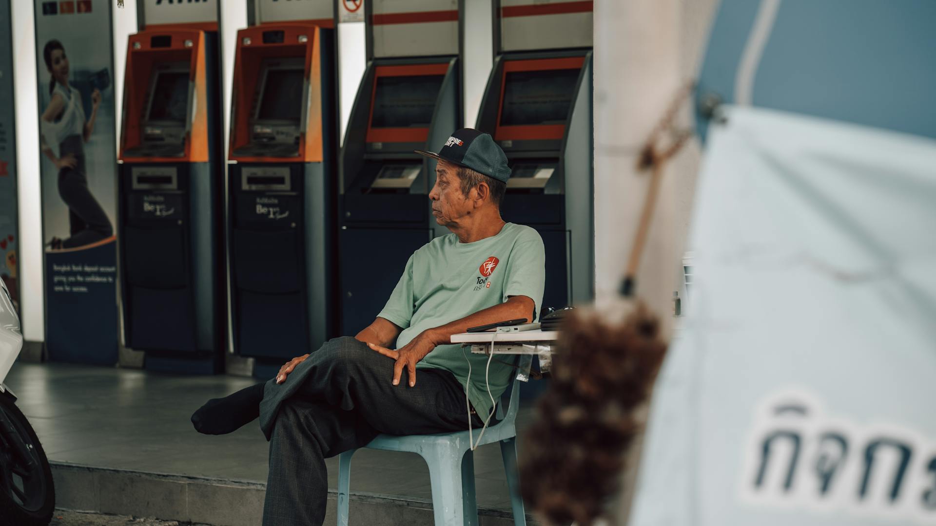 Elderly man sitting outside near ATM machines, looking away thoughtfully.