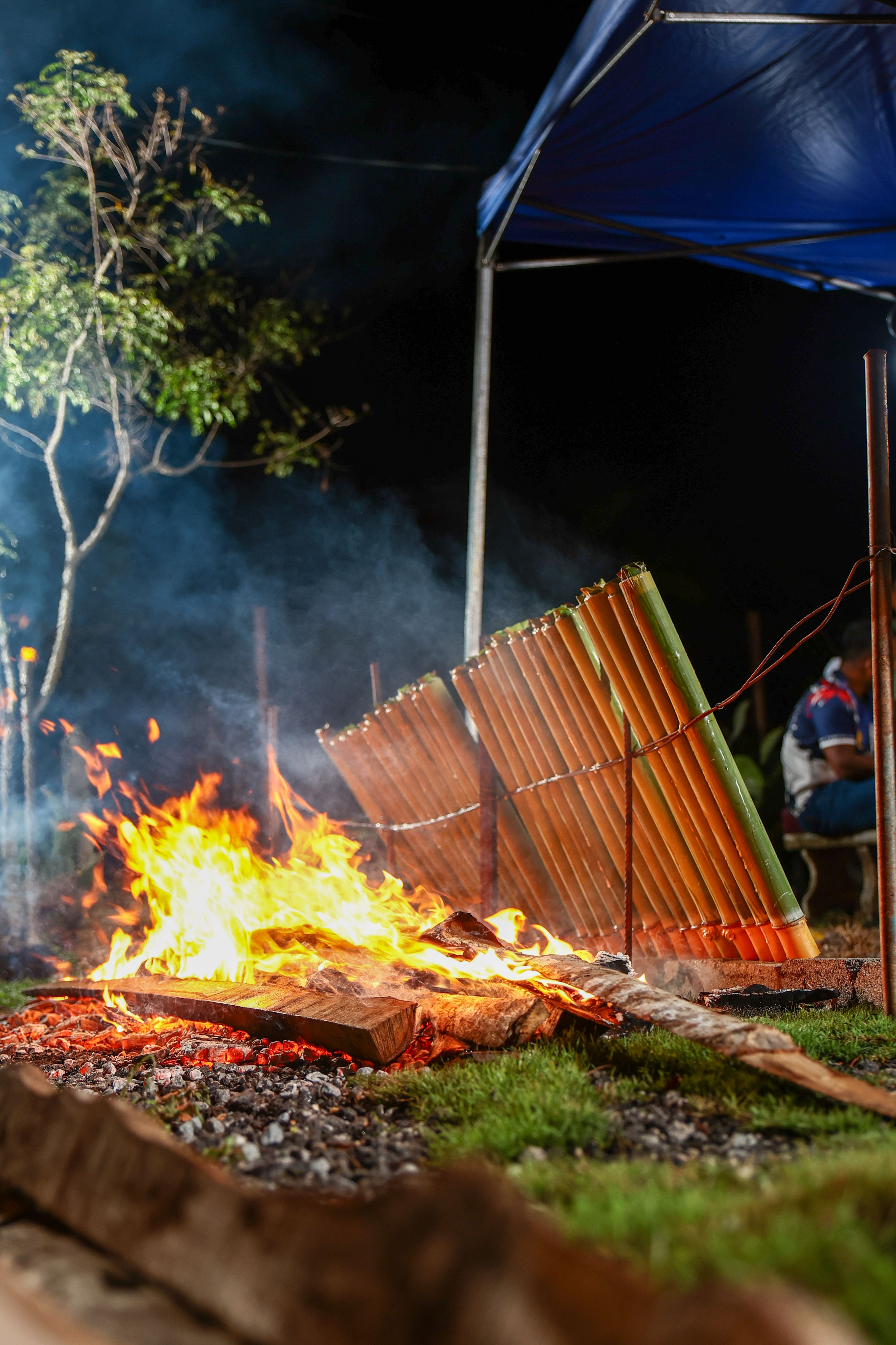 Bamboo filled with rice cooking over an open fire at night, showcasing traditional Malaysian outdoor cuisine.