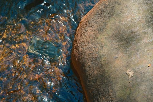 Free Sunlit water flows around a smooth rock in a tranquil natural stream setting. Stock Photo