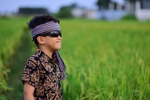 Free A young boy wearing sunglasses and a patterned shirt stands confidently in a vast green field under a clear sky. Stock Photo