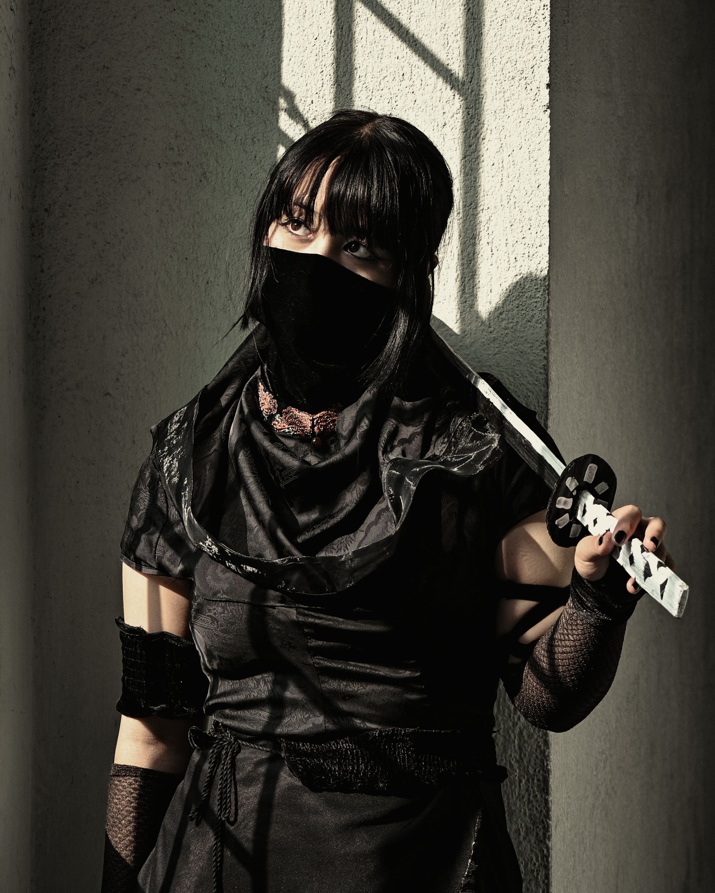 A woman dressed in black with a mask holds a sword in a shadow-filled space, exuding mystery and intensity.