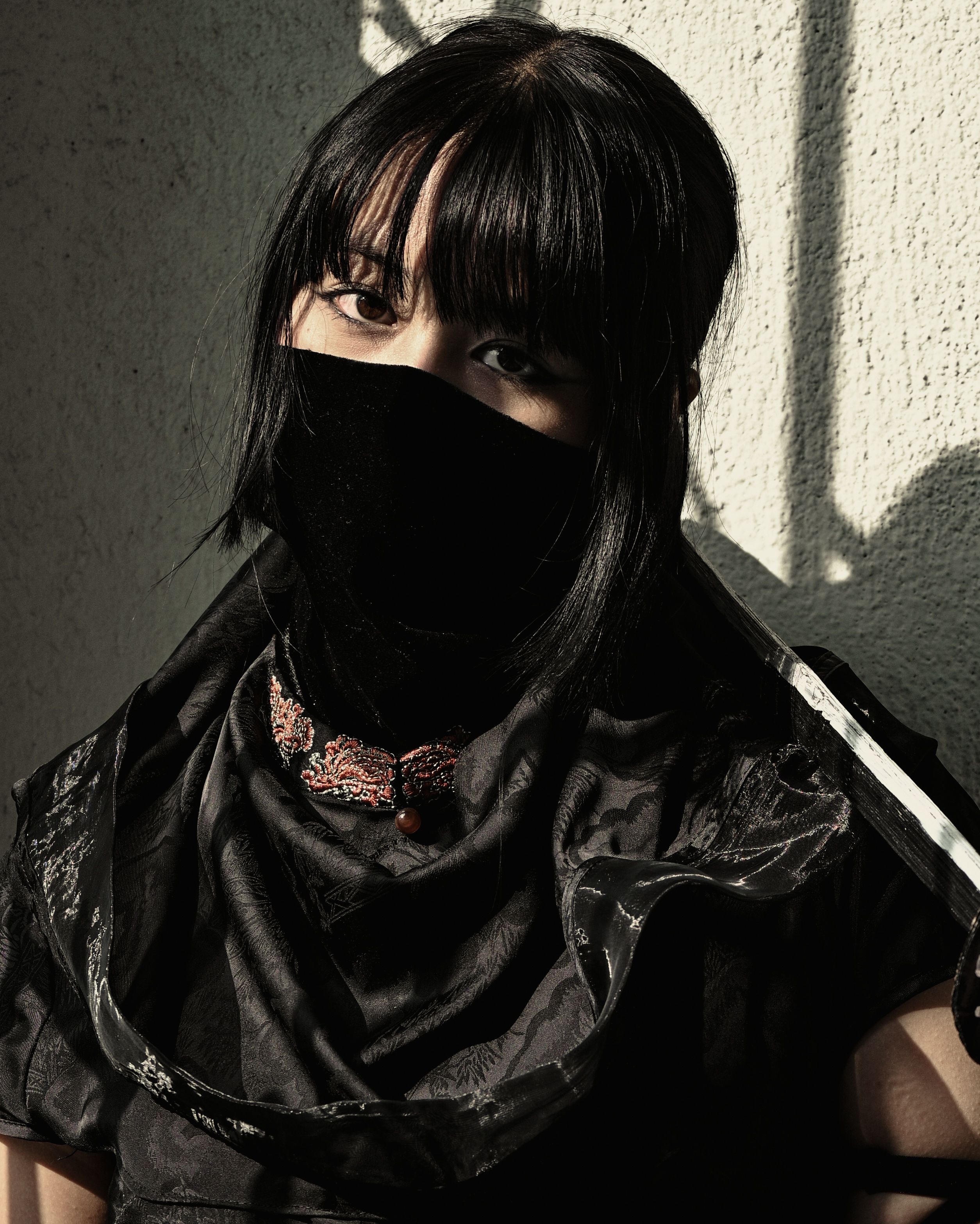 Intriguing portrait of a woman in a black mask, revealing only her intense eyes, set against a textured wall with dramatic lighting.
