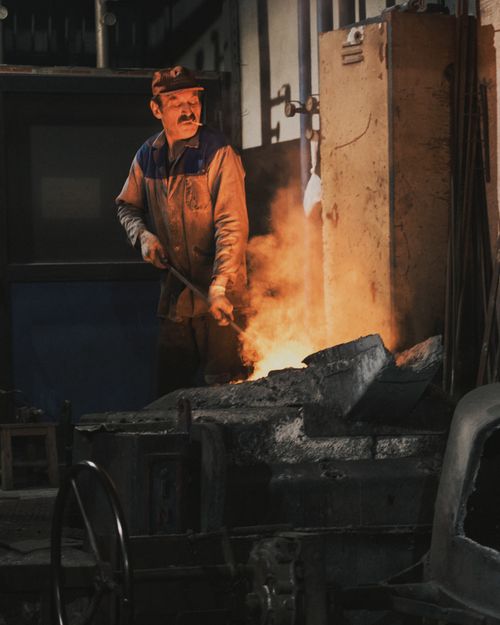 Free Blacksmith tending fire in a dimly lit forge, creating metalwork in an industrial setting. Stock Photo