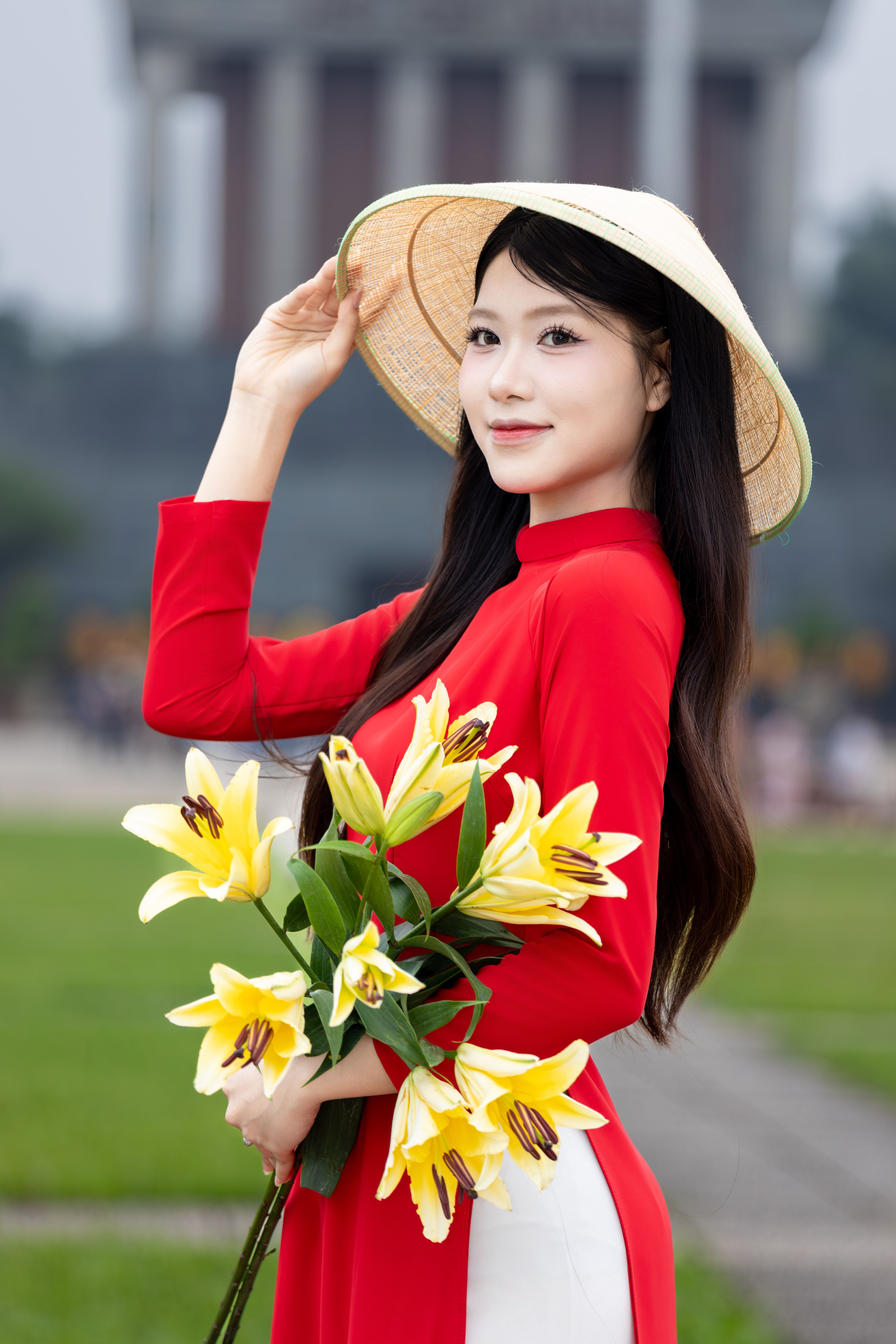 A Vietnamese woman wearing a traditional áo dài and conical hat, holding lilies outdoors in Hà Nội.