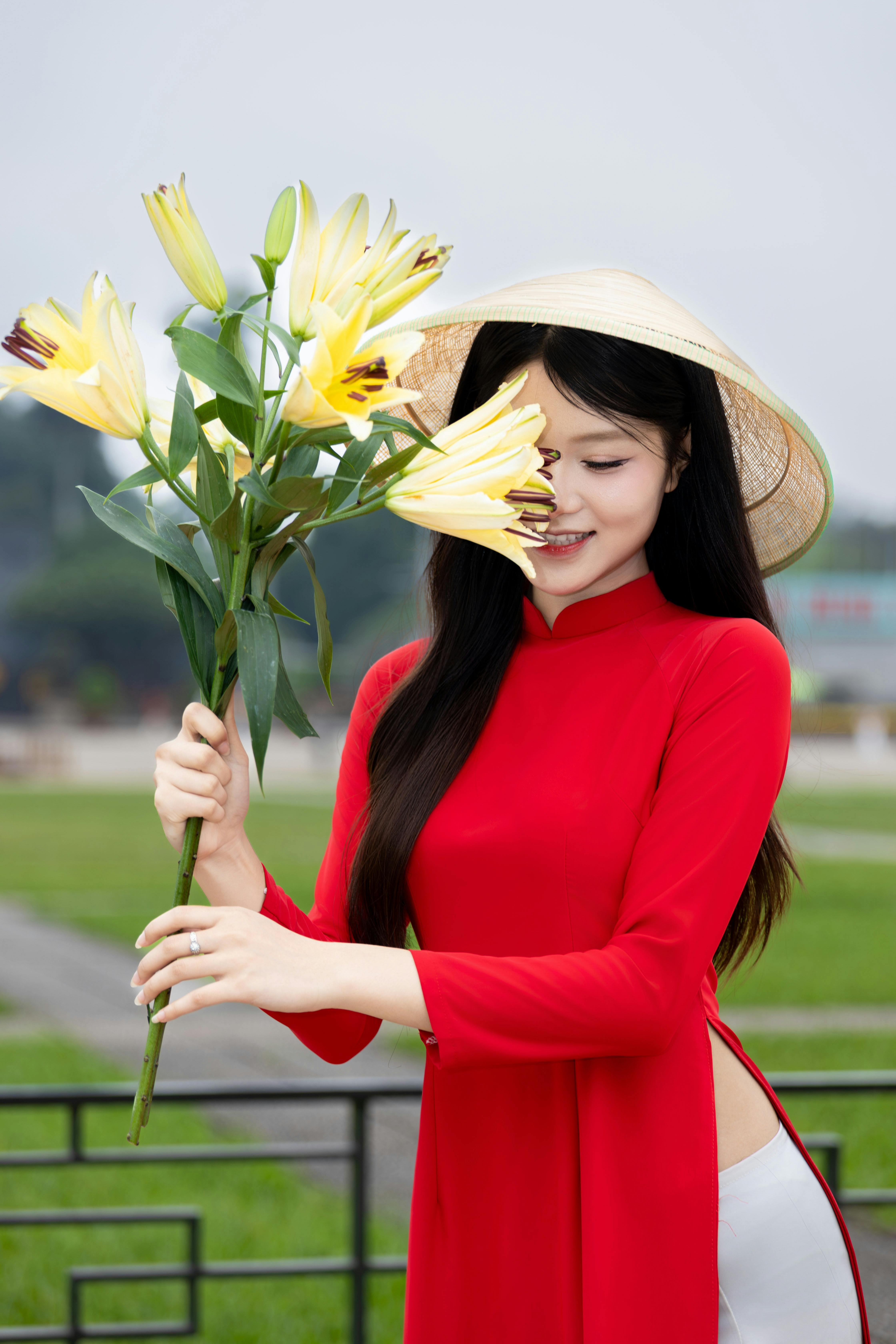 A woman wearing a red Ao Dai holding yellow lilies, outdoors in Hà Nội.
