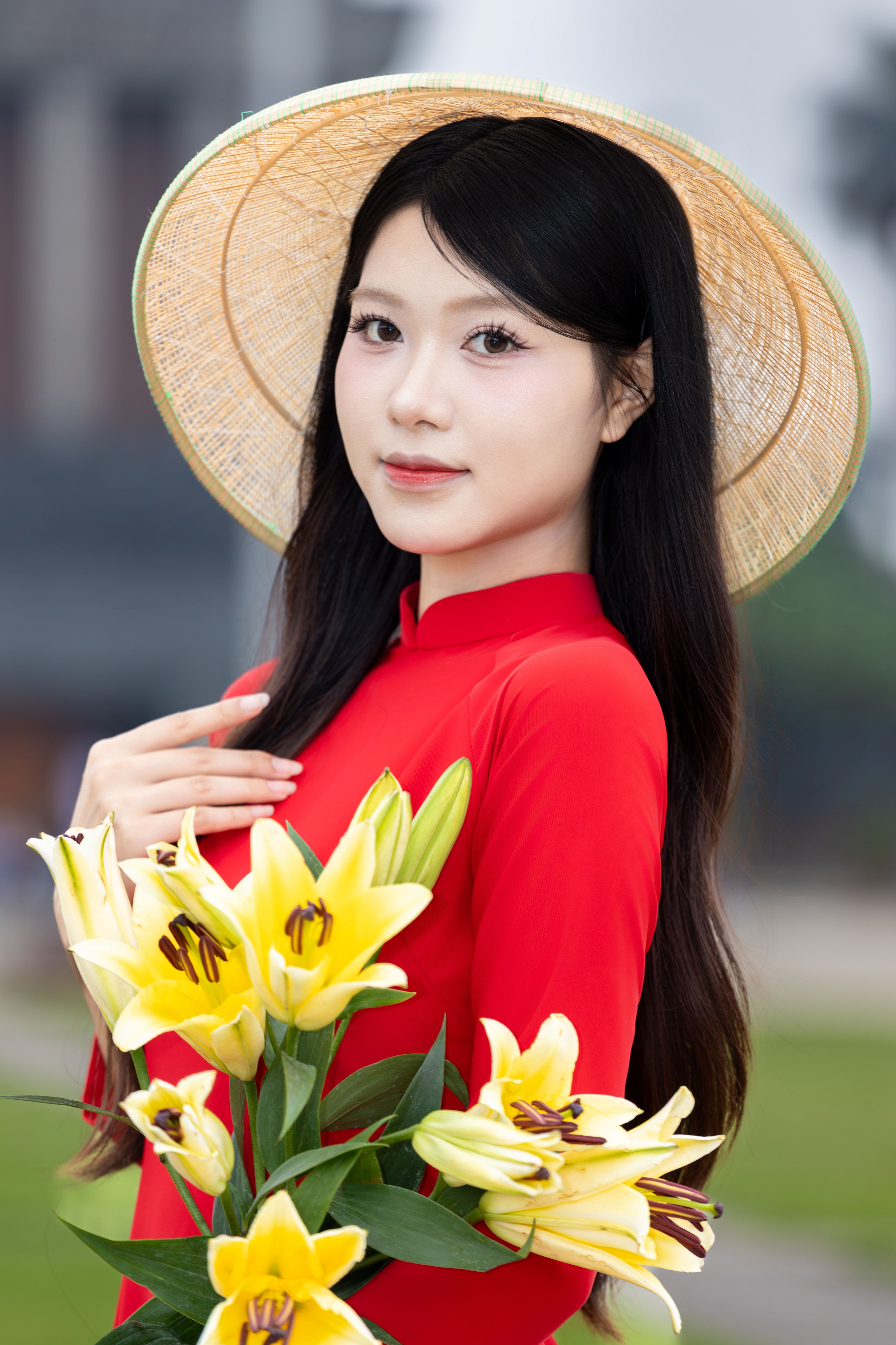 Young Vietnamese woman in red áo dài holding yellow lilies outdoors.